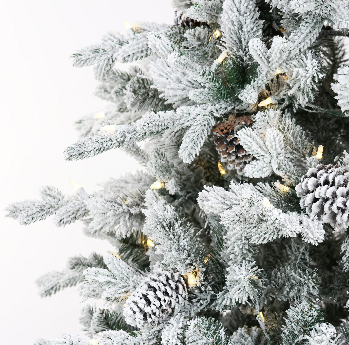 Close-up of a flocked Christmas tree with white snow-dusted branches, natural pinecones, and warm white string lights against a plain white background.