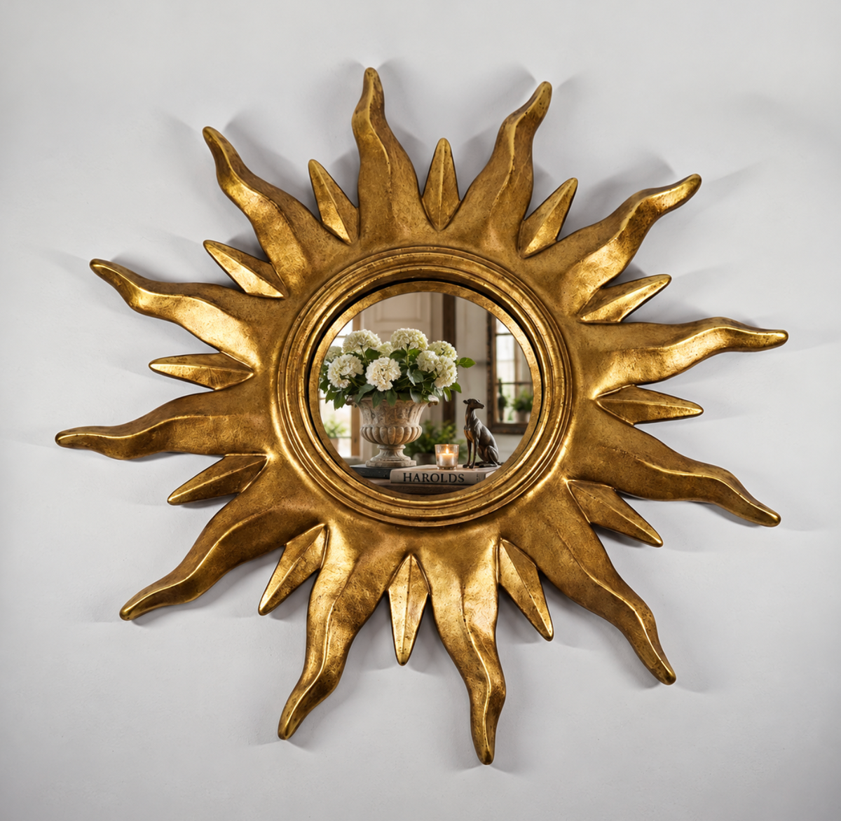 A gold sunburst wall mirror with a textured, sculptural frame, shown mounted on a light gray wall. The reflective center shows a decorative arrangement of white flowers and candles.