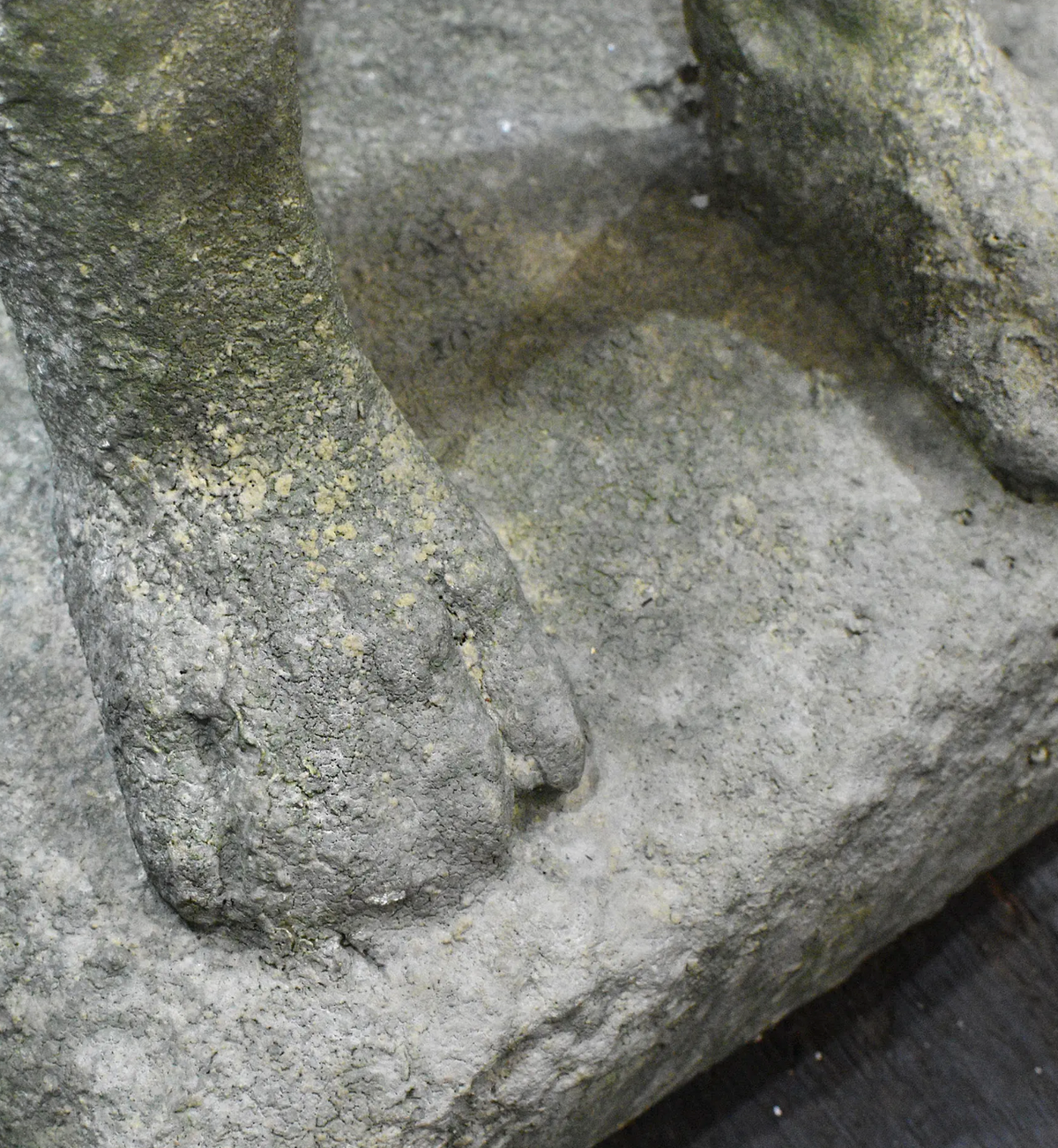 Close-up detail of a weathered stone sculpture or architectural feature, showing textured gray stone with mossy yellow-green patches and part of a carved foot or paw.