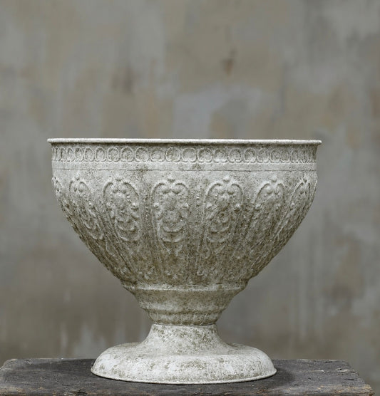 An ornate, vintage-style stone or concrete planter pot with embossed floral and scrollwork patterns, shown on a rustic wooden surface against a textured gray wall.