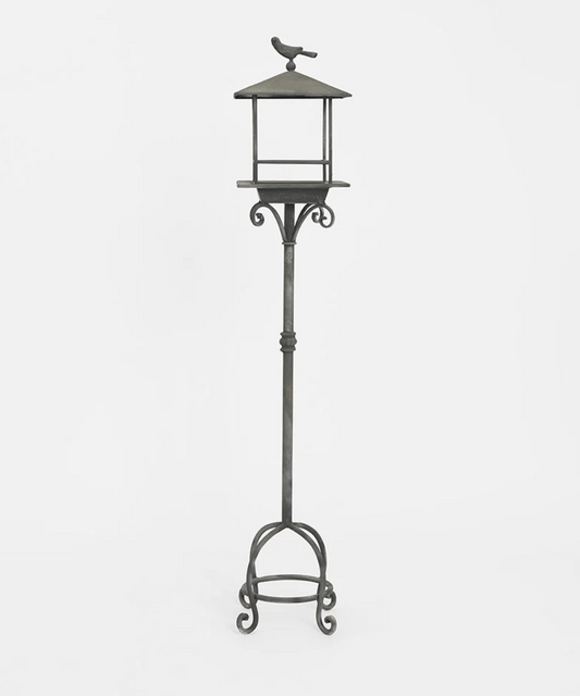 A tall wrought iron garden stake with a bird feeder on top, featuring a small bird perched at the top, set against a plain light gray background.