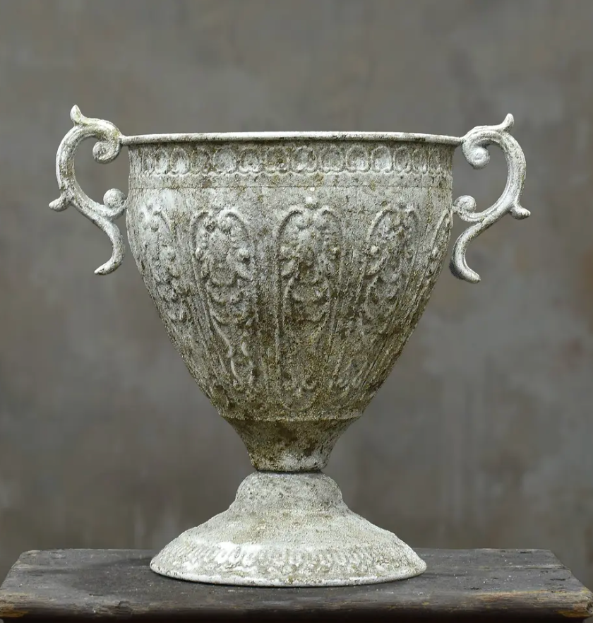 An ornate classical-style urn with two decorative handles, featuring embossed floral and scrollwork patterns, finished in a weathered silver-gray tone, displayed on a dark wooden surface against a textured gray background.