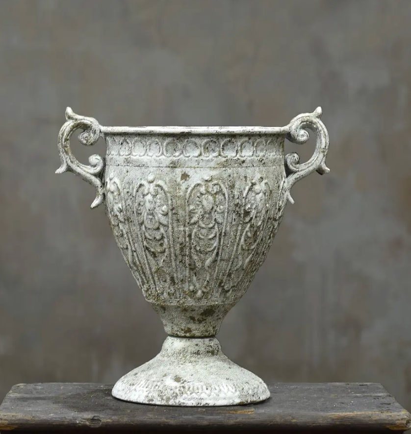 An ornate, vintage-style metal urn with a distressed, weathered silver finish, featuring decorative floral relief patterns and scrolled side handles, displayed on a dark wooden surface against a textured gray background.