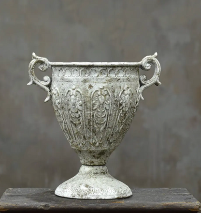 An ornate, vintage-style metal urn with a distressed, weathered silver finish, featuring decorative floral relief patterns and scrolled side handles, displayed on a dark wooden surface against a textured gray background.