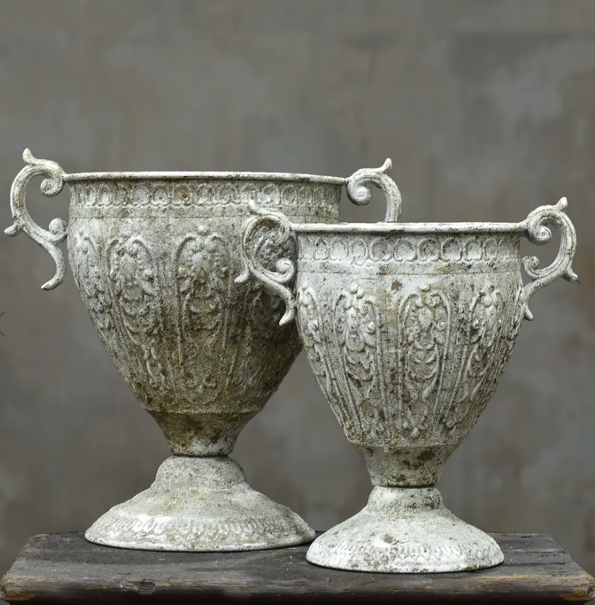 Two ornate vintage-style metal urns with decorative handles and embossed floral patterns, displayed on a rustic wooden surface against a neutral gray background.