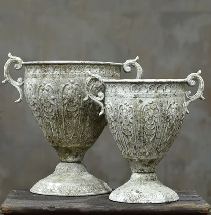 Two ornate vintage-style metal urns with decorative handles and embossed floral patterns, displayed on a rustic wooden surface against a neutral gray background.