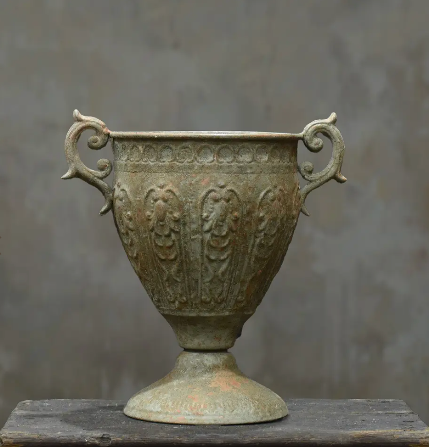 An antique bronze-colored metal cup with ornate handles and embossed floral patterns, displayed on a dark wooden surface against a textured gray background.