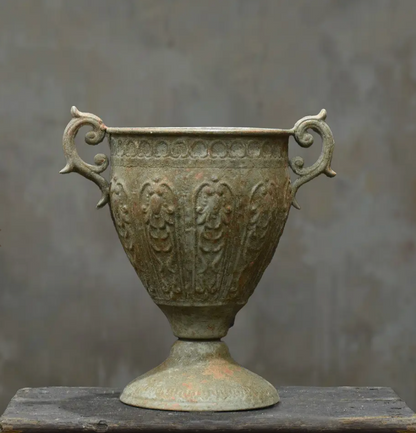 An antique bronze-colored metal cup with ornate handles and embossed floral patterns, displayed on a dark wooden surface against a textured gray background.