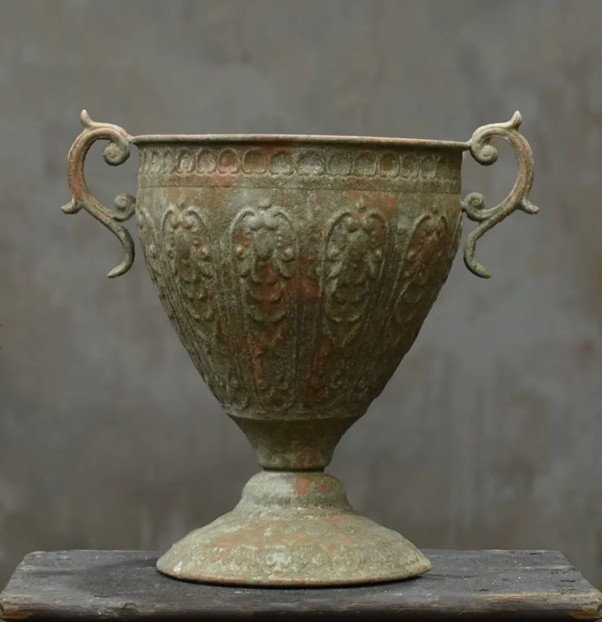 An antique bronze-colored metal urn with ornate handles and embossed floral patterns, displayed on a dark wooden surface against a textured gray background.