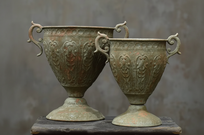 Two antique bronze urn-style vases with ornate handles and embossed floral relief patterns, displayed side by side on a dark wooden surface against a textured gray wall.