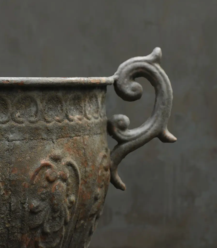 Close-up of an antique-style metal planter or urn with a weathered bronze and gray finish, featuring a decorative scroll handle against a dark gray background.