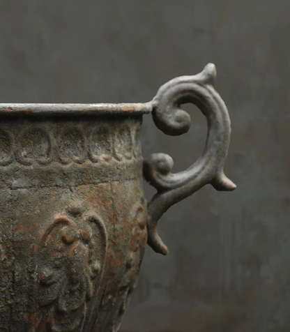 Close-up of an antique-style metal planter or urn with a weathered bronze and gray finish, featuring a decorative scroll handle against a dark gray background.