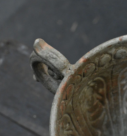 Close-up detail of an antique bronze or pewter vessel with a decorative handle and embossed floral pattern, showing weathered patina and rust-like discoloration against a dark gray background.
