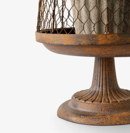 Close-up of a rustic pedestal table or planter base with a distressed brown and orange finish, featuring a fluted pedestal stem and a round tray, with a wire-mesh top container holding a tree trunk.