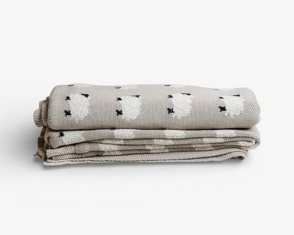A folded woven blanket in a light oatmeal beige color, featuring a pattern of white sheep with small black accents, set against a clean white background.