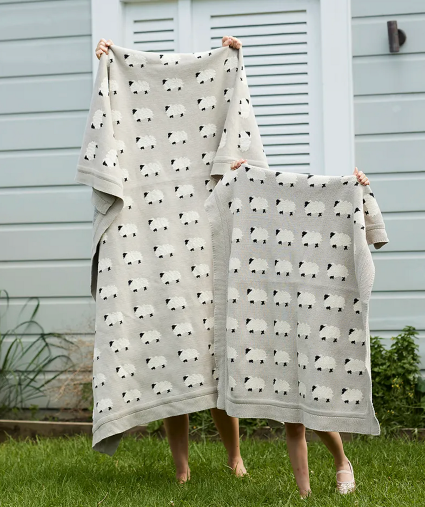 Two people standing outdoors on grass in front of a house, each holding up a light gray blanket or throw with an allover black-and-white panda print. The larger person holds a longer blanket, and the smaller person holds a shorter blanket.