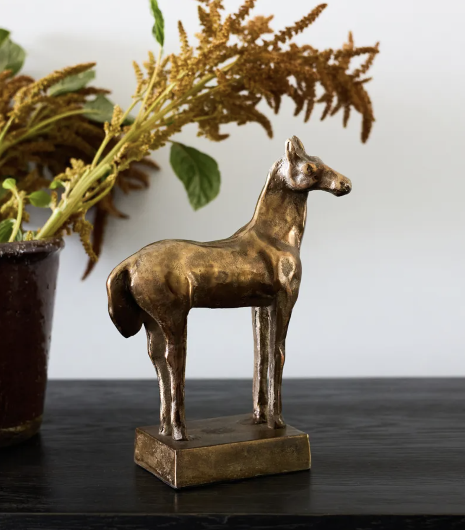 A small metallic horse figurine with a textured bronze finish, standing on a rectangular base on a dark tabletop with a blurred potted plant in the background.