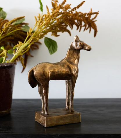 A small metallic horse figurine with a textured bronze finish, standing on a rectangular base on a dark tabletop with a blurred potted plant in the background.