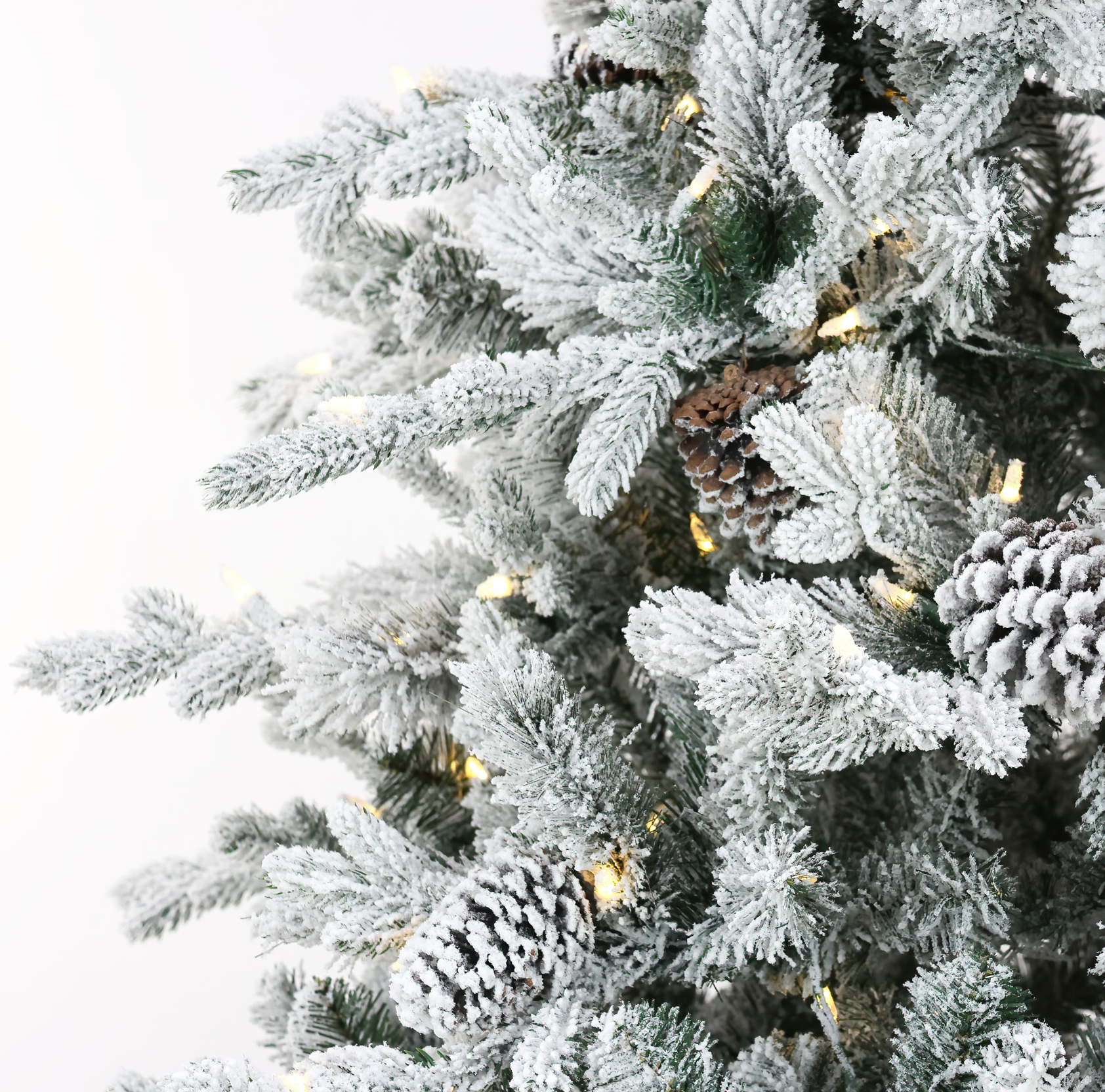 Close-up of a flocked Christmas tree with white snow-dusted branches, natural pinecones, and warm white string lights against a plain white background.