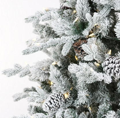 Close-up of a flocked Christmas tree with white snow-dusted branches, natural pinecones, and warm white string lights against a plain white background.