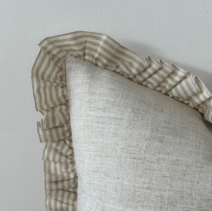 Close-up of a textured linen throw pillow with a beige and white striped ruffled fabric border, shown against a plain off-white wall.