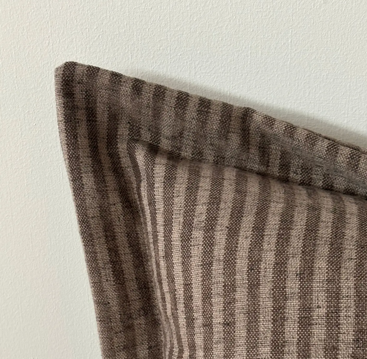 A close-up of a striped throw pillow corner in a brown and beige woven fabric, resting against a plain off-white wall.
