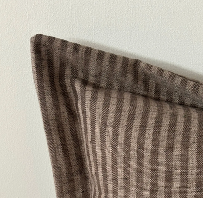 A close-up of a striped throw pillow corner in a brown and beige woven fabric, resting against a plain off-white wall.
