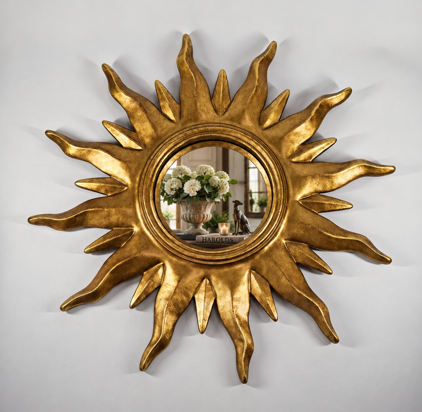 A gold sunburst wall mirror with a textured, sculptural frame, shown mounted on a light gray wall. The reflective center shows a decorative arrangement of white flowers and candles.