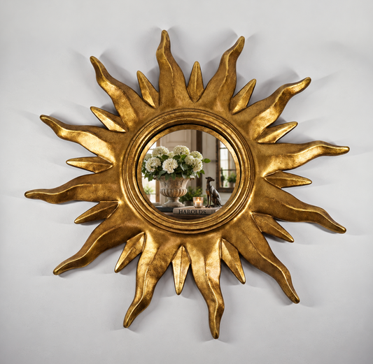 A gold sunburst wall mirror with a textured, sculptural frame, shown mounted on a light gray wall. The reflective center shows a decorative arrangement of white flowers and candles.