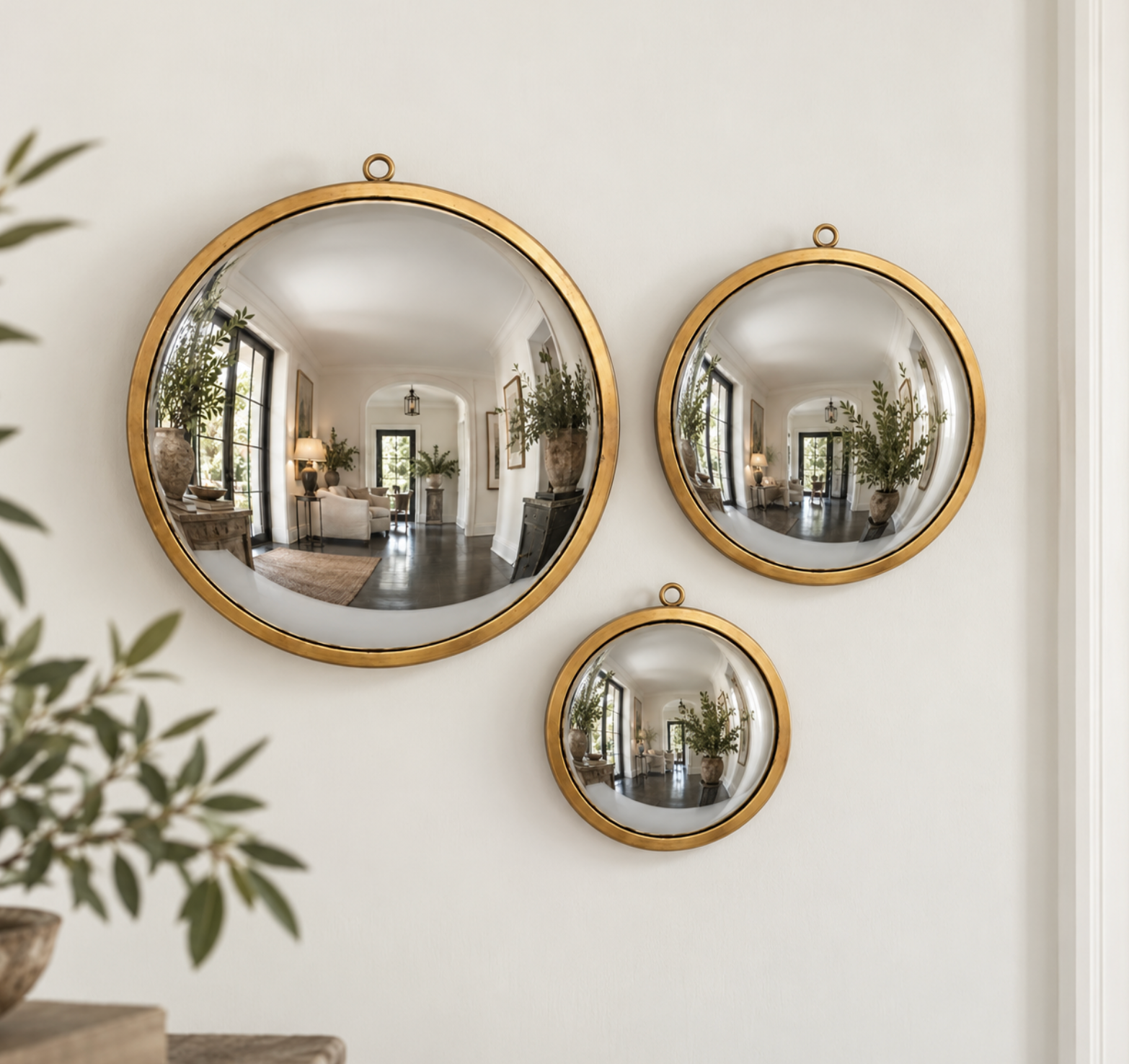 Three round convex security mirrors with gold frames of varying sizes, mounted on a white wall. The mirrors reflect a home interior with plants and furniture, and a blurred olive branch is visible in the foreground.