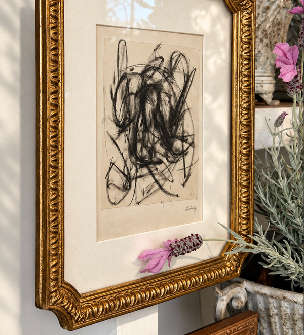 A framed abstract art print featuring black scribbled lines on beige paper, set in a white mat and an ornate gold frame, styled on a wall with dried pink flowers and a potted plant.