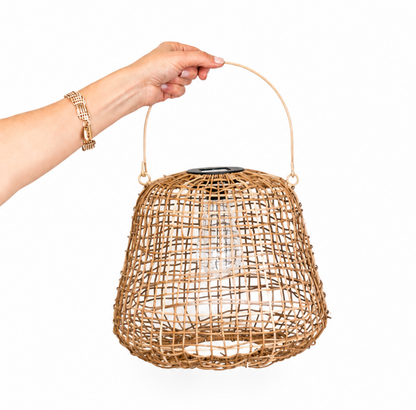 A hand holding a woven rattan basket-style pendant light with a curved handle and a visible clear bulb inside, against a plain white background.