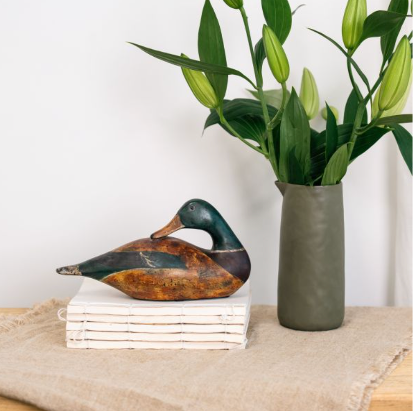 Small seated mallard duck sculpture with dark green head and brown body beside a vase