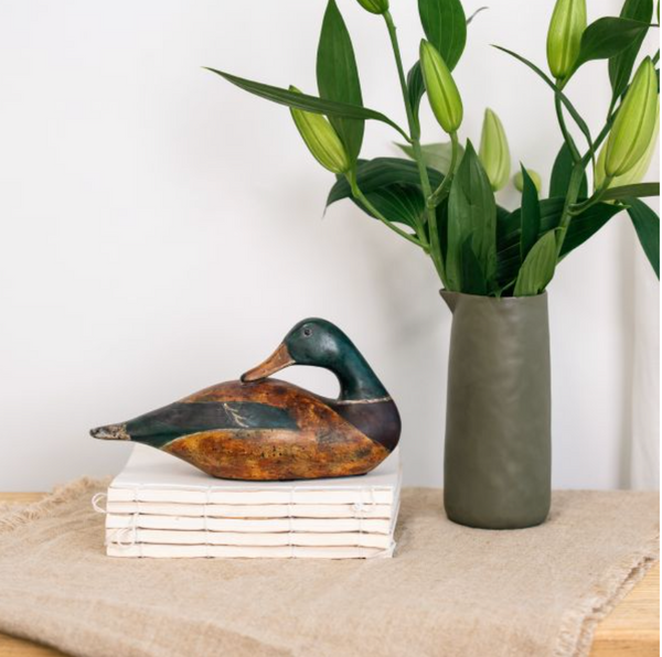 Small seated mallard duck sculpture with dark green head and brown body beside a vase