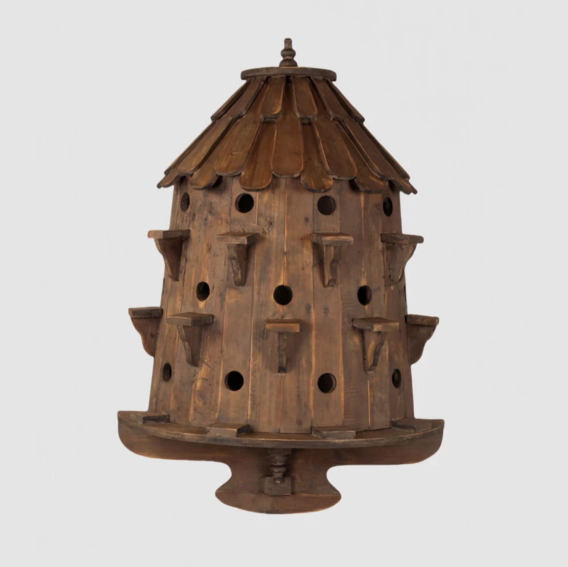 Brown wooden birdhouse with a shingled roof, rows of round openings, individual perches and a pedestal base.