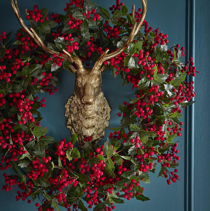 Holly and red berry wreath styled around a separate gold-coloured deer head against a dark blue wall