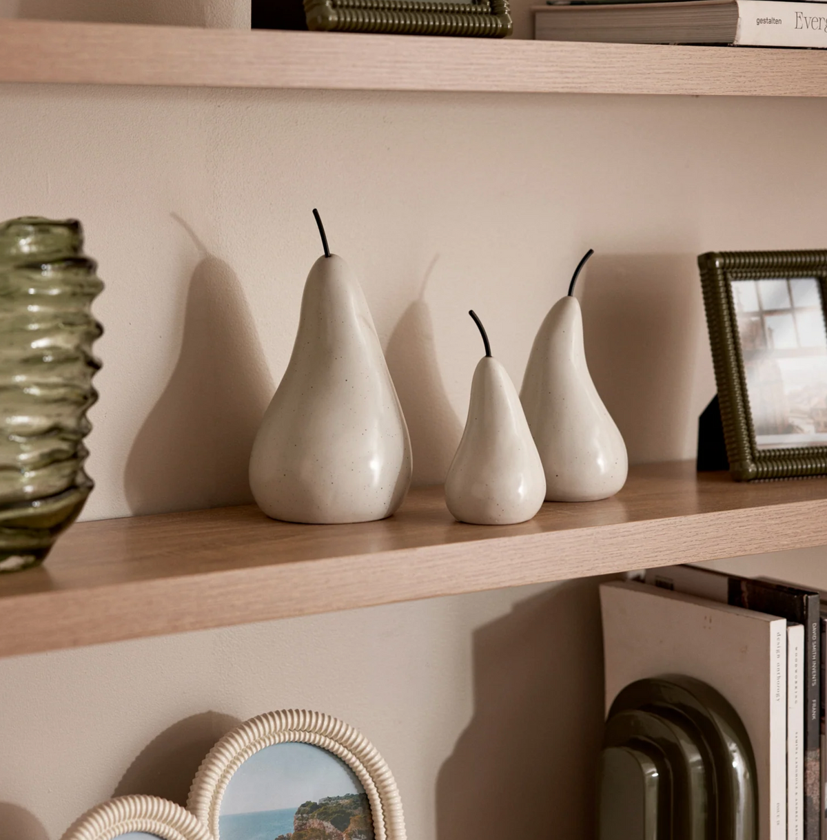 Off white ceramic pear sculptures arranged on a shelf beside a green vase and framed picture