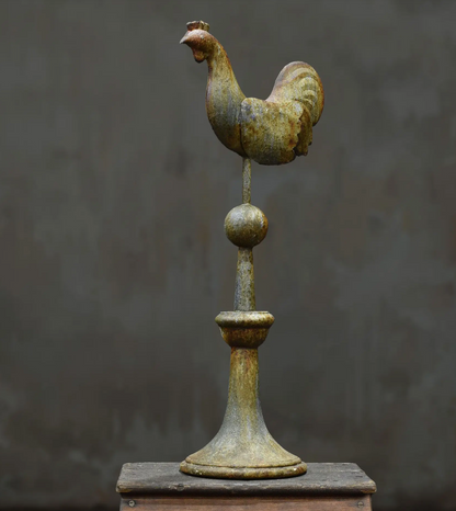 A vintage metal rooster weather vane with a distressed bronze and gold patina, mounted on a decorative post and displayed on a wooden block against a dark gray textured background.