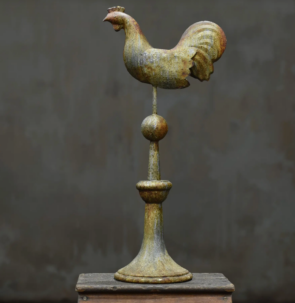 A rustic metal rooster weather vane mounted on a decorative post, displayed on a wooden surface against a dark textured background.