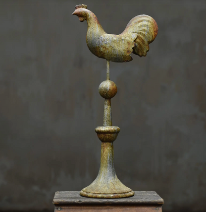 A rustic metal rooster weather vane mounted on a decorative post, displayed on a wooden surface against a dark textured background.