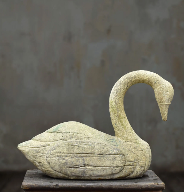 A carved stone swan sculpture with a textured, weathered finish, displayed on a rustic wooden pedestal against a neutral gray background.
