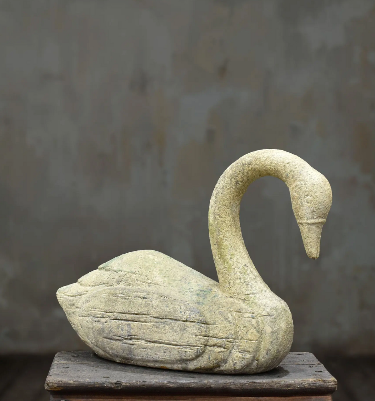 A carved stone swan sculpture in a natural light beige-gray hue, displayed on a rustic wooden pedestal against a textured gray background.