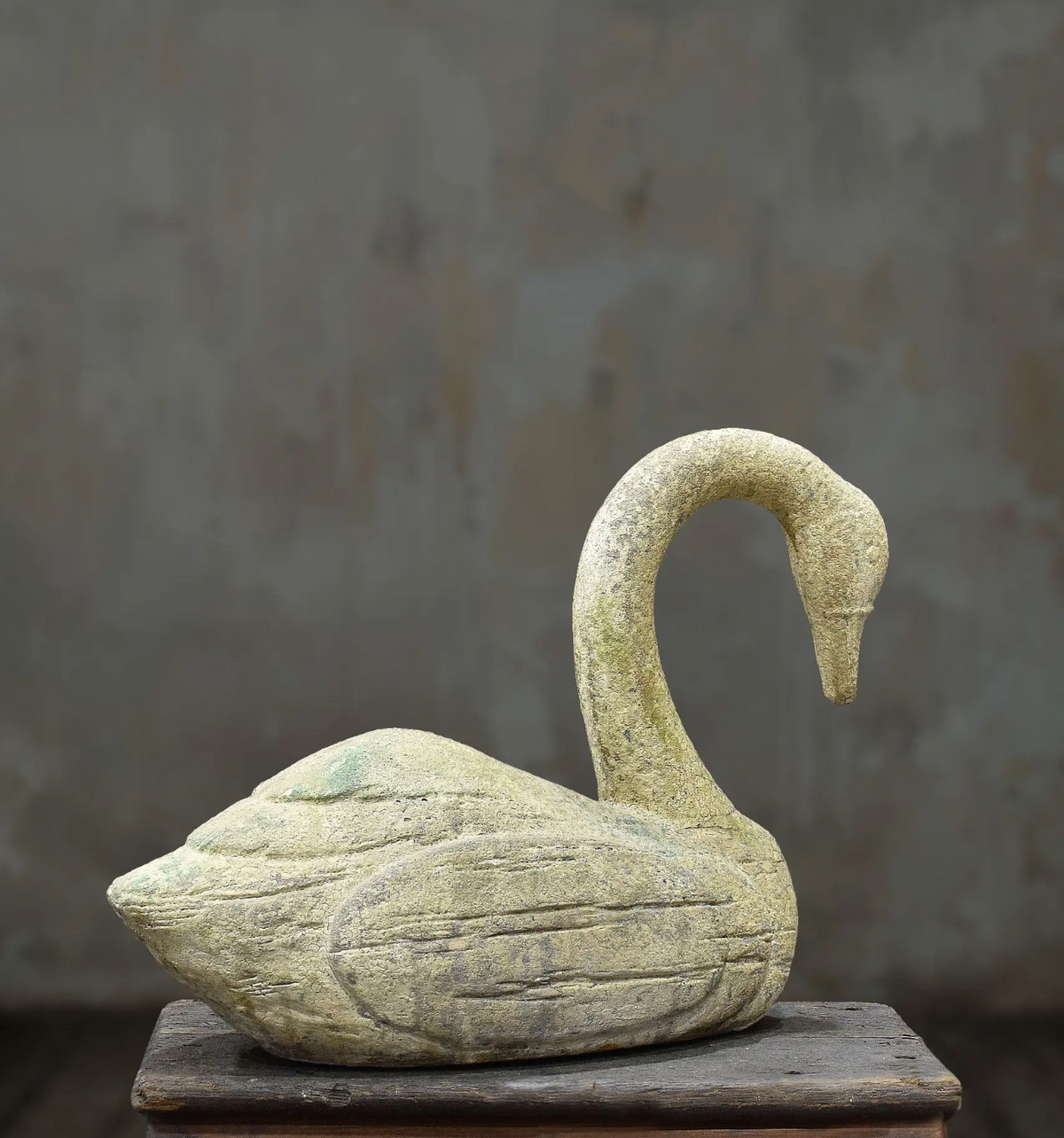 A carved stone swan sculpture with a textured, weathered finish, displayed on a rustic wooden pedestal against a neutral, textured gray background.