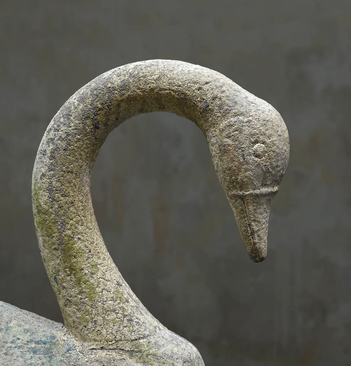 A close-up of a stone swan sculpture showing the textured neck and head in natural weathered gray and beige tones against a soft gray background.