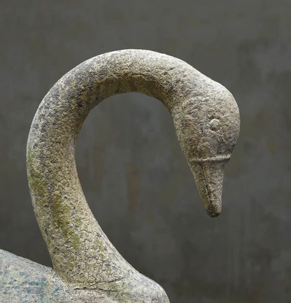 A close-up of a stone swan sculpture showing the textured neck and head in natural weathered gray and beige tones against a soft gray background.