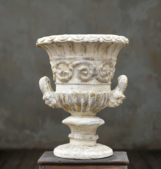 An ornate classical-style urn in a distressed off-white finish, displayed on a dark wooden pedestal against a textured gray background.