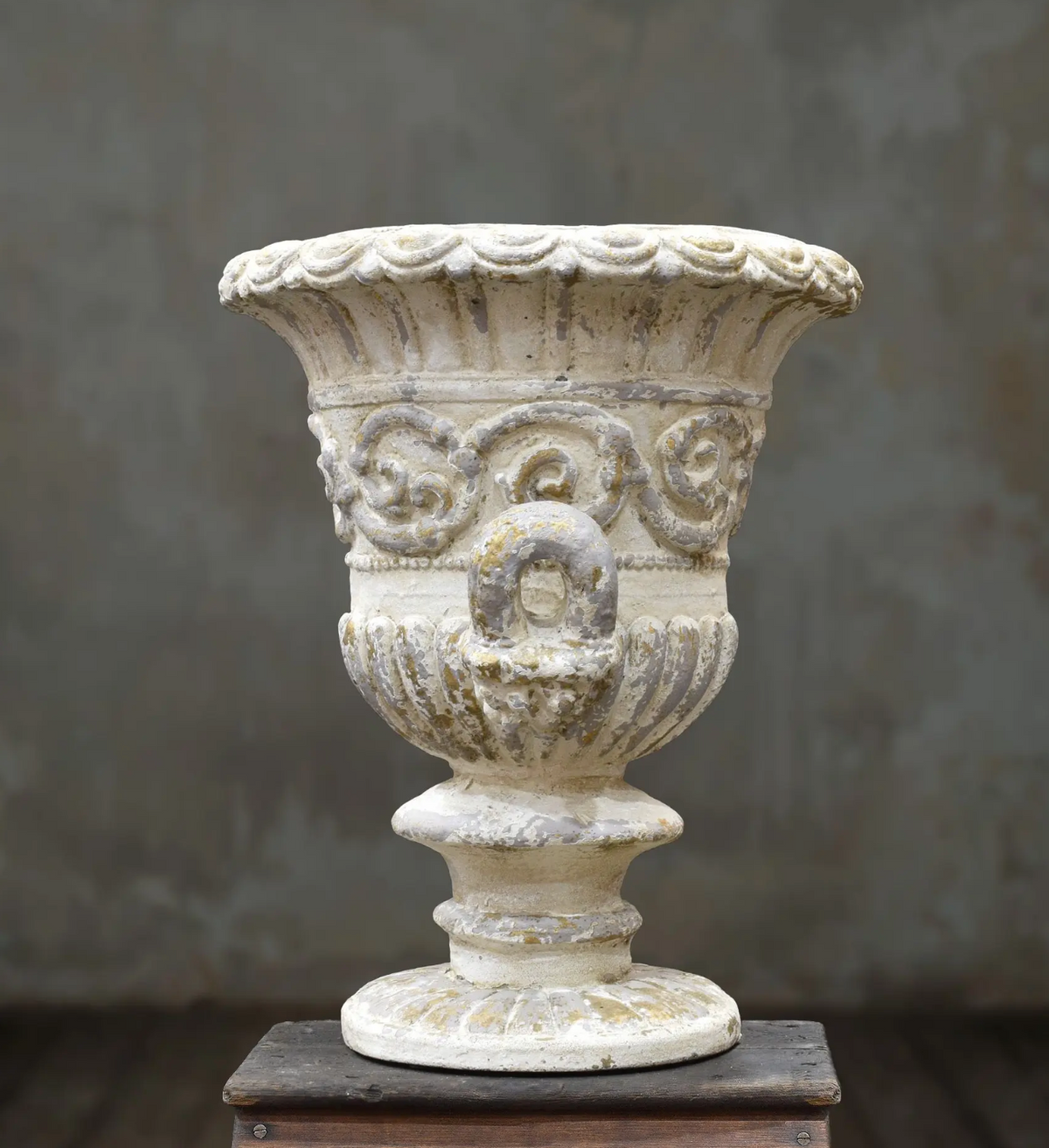 An ornate classical-style stone planter or urn in a weathered off-white finish, displayed on a dark wooden block against a textured gray studio backdrop.