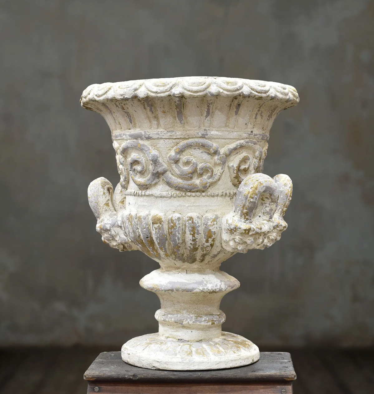 An ornate classical-style stone or concrete urn planter in a distressed off-white finish, displayed on a dark wooden block against a textured gray background.
